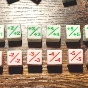 Pode incluir: Um conjunto de dados brancos com marcações verdes e vermelhas. Os dados são numerados de +1 a +6 e de -1 a -6. Os dados estão dispostos numa superfície de madeira, para jogos de tabuleiro.