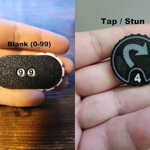 Pu&ograve; includere: Due contatori di gioco in bianco e nero. Il contatore a sinistra ha una forma ovale allungata con il testo Blank (0-99) e il numero 99. Il contatore a destra &egrave; circolare con un bordo seghettato, il testo Tap / Stun e il numero 4.