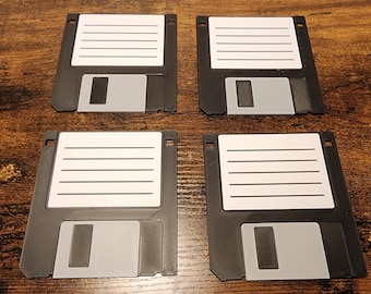 May include: Four black floppy disk coasters with white label areas and gray sliding tabs are arranged on a wooden surface. The coasters are square with rounded corners, evoking a retro computing aesthetic.
