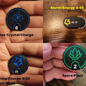 Pu&ograve; includere: Una collezione di quattro segnapunti di gioco circolari e uno oblungo, neri con superfici testurizzate. I segnapunti presentano icone e numeri colorati in blu, verde e giallo, incluse le etichette: Blue Crystal/Charge, Storm/Energy 0-99, Lightning/Charge 0-20 e Spore/Plant.