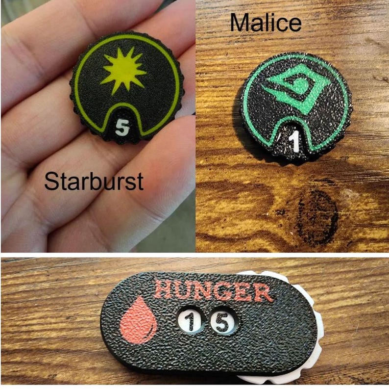 Pu&ograve; includere: Tre gettoni da gioco neri testurizzati. Un gettone rotondo presenta una stella gialla e il numero 5. Un altro gettone rotondo mostra un simbolo verde e il numero 1 etichettato come Malice. Un gettone ovale mostra una goccia rossa, la parola HUNGER e il numero 15.