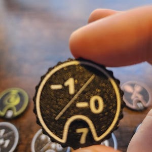 May include: A circular black game token with a textured surface featuring a white printed design. The design includes the numbers -1 and 0 separated by a diagonal line, with the number 7 positioned inside a small arch at the bottom edge.