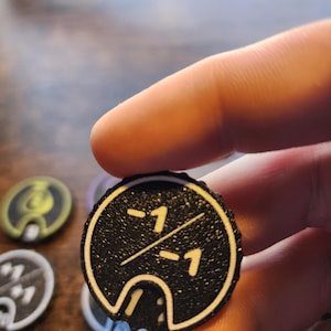 May include: A circular black game token with a textured surface featuring a white border and the white text -1 over -1 separated by a diagonal line. The token has a small cutout at the bottom edge and is held against a blurred background.