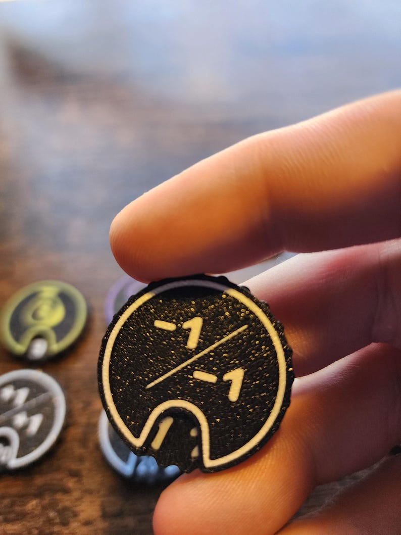 May include: A circular black game token with a textured surface featuring a white border and the white text -1 over -1 separated by a diagonal line. The token has a small cutout at the bottom edge and is held against a blurred background.