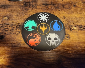 May include: A black circular mat featuring six colorful symbols, including a sun, a tree, a flame, a drop, a skull, and a lightning bolt. The symbols are arranged around a central point. The mat is on a wooden surface.