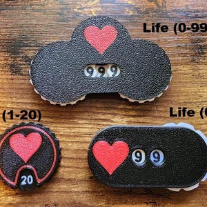 Pu&ograve; includere: Tre contatori di punti vita neri con icone a forma di cuore rosso e numeri bianchi. Il contatore superiore mostra 999, quello circolare in basso a sinistra mostra 20 e quello ovale in basso a destra mostra 99. Etichette: Life (0-999), Life (1-20), Life (0-99).