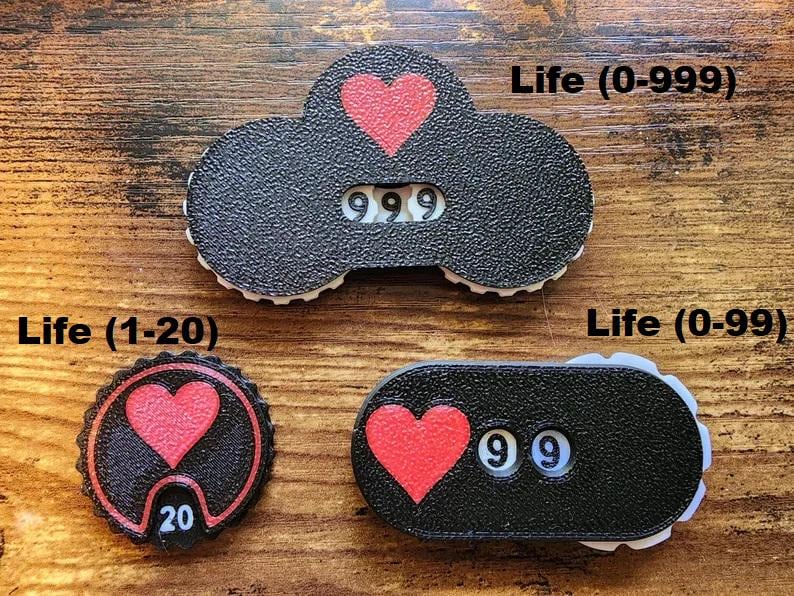 Pu&ograve; includere: Tre contatori di punti vita neri con icone a forma di cuore rosso e numeri bianchi. Il contatore superiore mostra 999, quello circolare in basso a sinistra mostra 20 e quello ovale in basso a destra mostra 99. Etichette: Life (0-999), Life (1-20), Life (0-99).