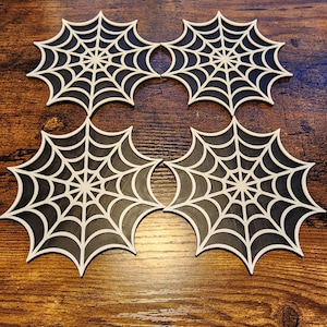 May include: Four spiderweb-shaped coasters. Each coaster features a black web design with white outlines. The coasters are arranged in a square formation on a wooden surface, perfect for Halloween or spooky decor.