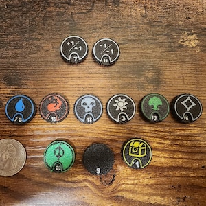 May include: A collection of circular black game tokens featuring various white, blue, red, green, and yellow icons including symbols for water, fire, skulls, suns, trees, and treasure chests, alongside a silver US quarter for scale reference.