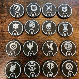 May include: Sixteen circular black tokens with white borders and white icons. Each token features a unique symbol in the center and a number at the bottom, ranging from 1 to 11. The symbols include skulls, shields, swords, towers, and various abstract shapes.