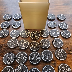 Puede incluir: Una colección de fichas de juego en blanco y negro con una caja dorada. Las fichas son redondas con un borde dentado y presentan símbolos y números blancos. La caja es rectangular y tiene un acabado dorado texturizado. Las fichas están dispuestas sobre una superficie de madera.