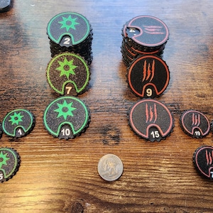 May include: A selection of black and green game tokens with starburst designs and numbers, and black and red tokens with claw marks and numbers. A US quarter is also present.