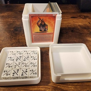 Puede incluir: Un contenedor de almacenamiento de plástico blanco para cartas y dados con una pila de cartas y dados. El contenedor tiene forma cuadrada con tapa y un compartimento para cartas. Los dados son blancos con marcas negras.