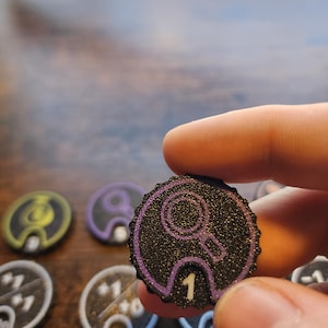 Pu&ograve; includere: Un gettone da gioco circolare nero con una superficie testurizzata e scintillante e un bordo viola. Il gettone presenta un'icona a forma di lente d'ingrandimento viola in alto e il numero 1 bianco stampato al centro in basso in una tacca.