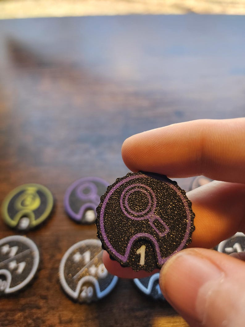 Pu&ograve; includere: Un gettone da gioco circolare nero con una superficie testurizzata e scintillante e un bordo viola. Il gettone presenta un'icona a forma di lente d'ingrandimento viola in alto e il numero 1 bianco stampato al centro in basso in una tacca.