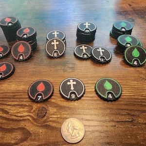 Può includere: Una collezione di gettoni da gioco circolari neri con icone rosse, bianche e verdi di gocce di sangue e croci, ciascuno contrassegnato da un valore numerico. Una moneta da un quarto di dollaro statunitense è posta in basso come riferimento.