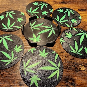 May include: Set of seven round black coasters with a green marijuana leaf design. The coasters are made of a textured material and have a black base.
