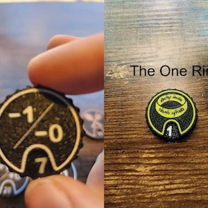 Pu&ograve; includere: Un gettone circolare nero con superficie testurizzata e dettagli stampati in giallo. Un lato mostra i numeri -1, -0 e 7. L'altro lato presenta un grafico di un anello con il testo The One Ring e il numero 1 sul bordo inferiore.