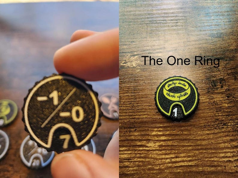 Pu&ograve; includere: Un gettone circolare nero con superficie testurizzata e dettagli stampati in giallo. Un lato mostra i numeri -1, -0 e 7. L'altro lato presenta un grafico di un anello con il testo The One Ring e il numero 1 sul bordo inferiore.