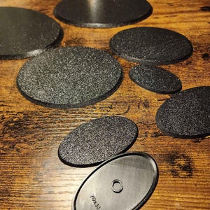 May include: A selection of black, textured coasters in different shapes, including round and oval. The coasters have a slightly rough surface and are arranged on a wooden surface. One coaster has the text "16x32".