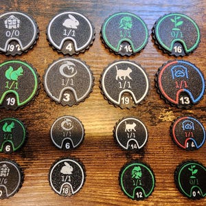 May include: A collection of sixteen circular black game tokens with scalloped edges, featuring various white, green, blue, or red icons of animals and plants, along with printed numerical values like 1/1, 0/0, 0/1, 19, 4, 16, 3, 13, 6, 14, 10, 18, and 12.