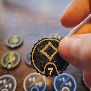 Pu&ograve; includere: Un gettone circolare nero con bordo smerlato caratterizzato da un simbolo a rombo dorato in rilievo e il numero 7 al centro. Il gettone ha una superficie ruvida e fa parte di una collezione di segnapunti di gioco simili con vari numeri.