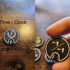 Pu&ograve; includere: Un gettone nero circolare con bordo bianco e un'icona bianca di un orologio con una freccia in senso antiorario sopra il numero 7, etichettato Time / Clock. Un altro gettone nero presenta una linea diagonale bianca che separa due valori -1.