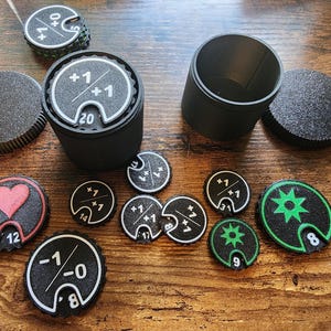 Peut inclure: Gobelet noir pour dés de jeu avec couvercle et divers jetons de jeu. Les jetons comportent des chiffres, des symboles et des valeurs comme +1, -1 et des symboles de cœur. Les jetons sont noirs avec des accents blancs et verts. Le gobelet mesure environ 10 cm de haut.