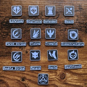 May include: A collection of small, square, black and white game tokens with various symbols and text. The tokens include phrases like "Lifelink," "Vigilance," "Defender," and "Reach." The tokens are arranged on a wooden surface.