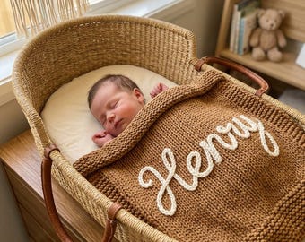 May include: A woven wicker Moses basket with brown leather handles containing a sleeping infant covered by a thick, textured, rust-colored knit blanket. The blanket features the name Henry embroidered in white cursive yarn lettering.