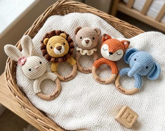 May include: Five crocheted animal rattles on wooden rings, featuring a rabbit, lion, bear, fox, and elephant. Each wooden ring is engraved with a name: Sophie, James, Teddy, Oliver, and Lucas. A wooden block with the letter B sits nearby.