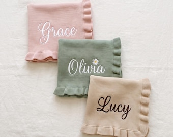 May include: Three knitted blankets with ruffled edges in dusty pink, sage green, and beige. The pink blanket features the name Grace in white script, the green blanket features Olivia with a daisy, and the beige blanket features Lucy in brown.