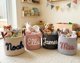 May include: Four round, ribbed fabric storage baskets with brown leather handles. Each basket features a personalized name in raised, stitched lettering: Noah in dark blue, Ella in white, James in white, and Mia in pink, filled with toys.