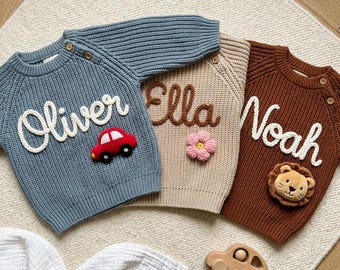 May include: Three long-sleeve knit sweaters in blue, beige, and brown. The blue sweater features the name Oliver and a red car appliqué. The beige sweater displays the name Ella and a pink flower. The brown sweater shows the name Noah and a lion face.