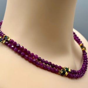 May include: A double-strand necklace featuring faceted, deep magenta-colored beads. The strands are accented at intervals with small, gold-toned metal spacers that incorporate dark green, textured cylindrical elements between the beads.