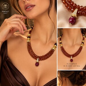 May include: A multi-strand necklace featuring deep red faceted gemstone beads, gold-toned metal spacers, and a central pendant with a round red stone and clear crystals. The necklace is displayed on a neck and includes text overlays.
