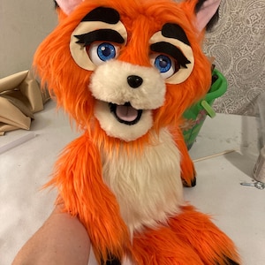 May include: A vibrant orange fox fursuit head with large blue eyes, black accents, and a white muzzle. The fursuit features a friendly expression and is made of soft, plush material. The fox has black-tipped ears and a fluffy orange body.