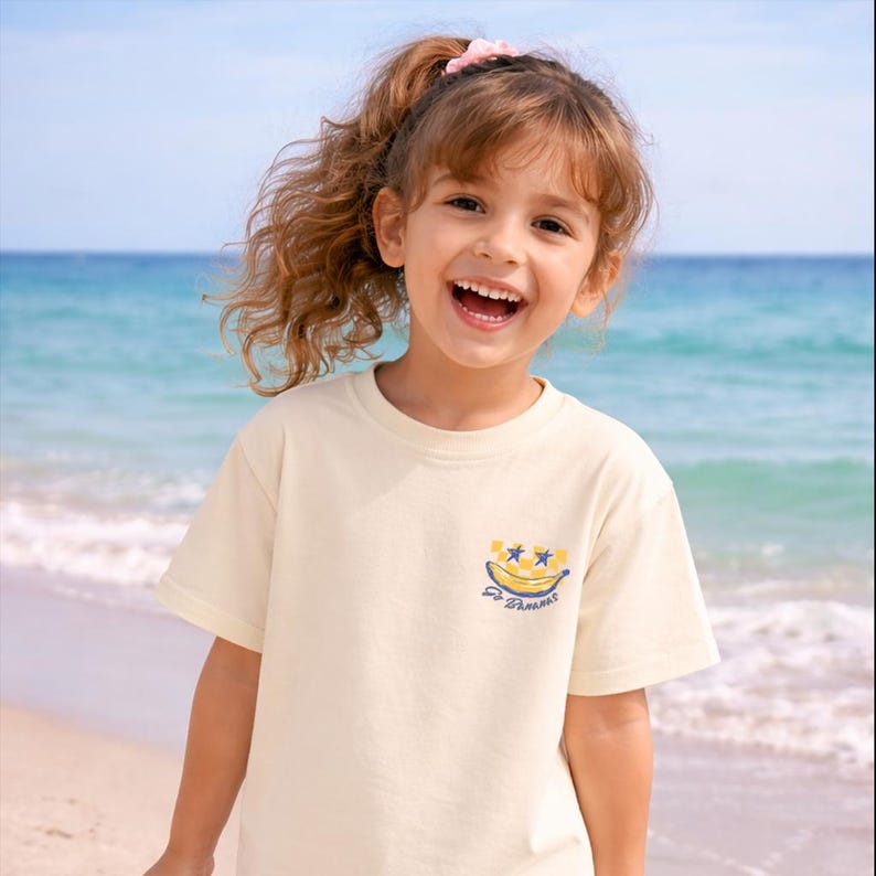 May include: A young girl smiles broadly, wearing a cream-colored t-shirt with a banana graphic and the text "Go Banana". The background features a beach with blue ocean water and a clear sky.