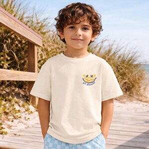 May include: Cream-colored t-shirt with a banana graphic and the text "So Bananas". The shirt is short-sleeved and has a crew neck. The child is wearing blue and white checkered shorts.