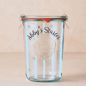 May include: A clear glass jar with a red lid and metal clasps. The jar is decorated with light blue vertical stripes, small white daisies, and the text "Abby's Starter". The jar has a Weck logo.