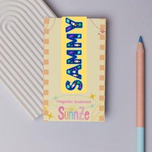 May include: A yellow magnetic bookmark featuring the name "SAMMY" in blue, space-themed letters. The bookmark is on a light yellow book with the text "magnetic bookmark" and the brand name "Sunnze". A blue pencil is on the right.