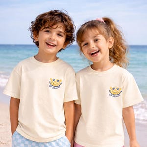 May include: Two children wearing cream-colored t-shirts with a banana graphic. The graphic includes a smiling banana, two stars, and the text "So Bananas" in blue. The children are smiling in front of a beach background.