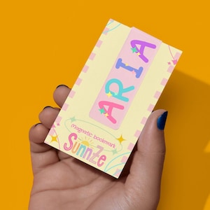 May include: A magnetic bookmark with the name "ARIA" in purple, blue, and pink letters, decorated with stars. The bookmark also features the text "magnetic bookmark Sunnze" in a playful font, set against a yellow background.