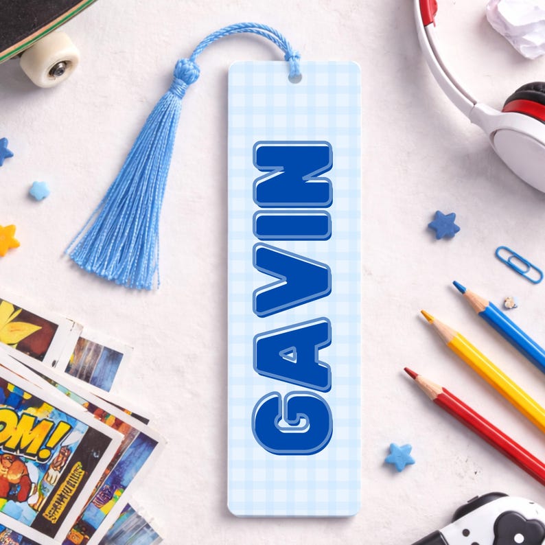 May include: A personalised bookmark featuring the name "GAVIN" in bold blue lettering. The bookmark has a light blue gingham pattern and a matching blue tassel. Pencils, a skateboard, and headphones are in the background.