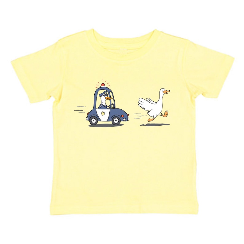 May include: A yellow short-sleeved t-shirt featuring a cartoon illustration of a goose running away from a police car driven by a goose.