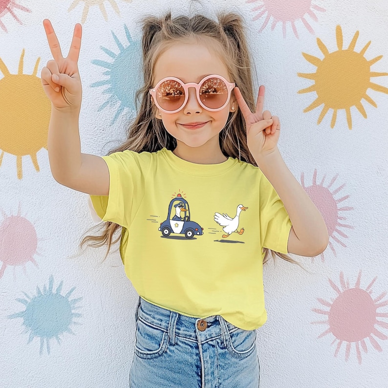 May include: A young girl wearing pink sunglasses and a yellow t-shirt with a cartoon illustration of a police car chasing a goose. The girl is smiling and has her hair pulled back in a ponytail.