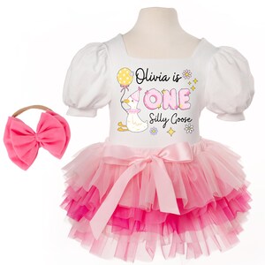 May include: A white and pink baby outfit with a matching pink bow headband. The outfit features a white top with puff sleeves and a tutu skirt in shades of pink. Text on the top reads "Olivia is ONE Silly Goose" with a cartoon goose illustration.