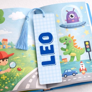 May include: A light blue bookmark with the name "LEO" in bold blue letters. The bookmark has a light blue tassel and is placed in a children's book with colourful illustrations of a dinosaur, alien, and rocket ship.