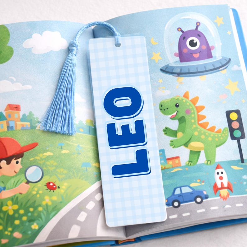 May include: A light blue bookmark with the name "LEO" in bold blue letters. The bookmark has a light blue tassel and is placed in a children's book with colourful illustrations of a dinosaur, alien, and rocket ship.