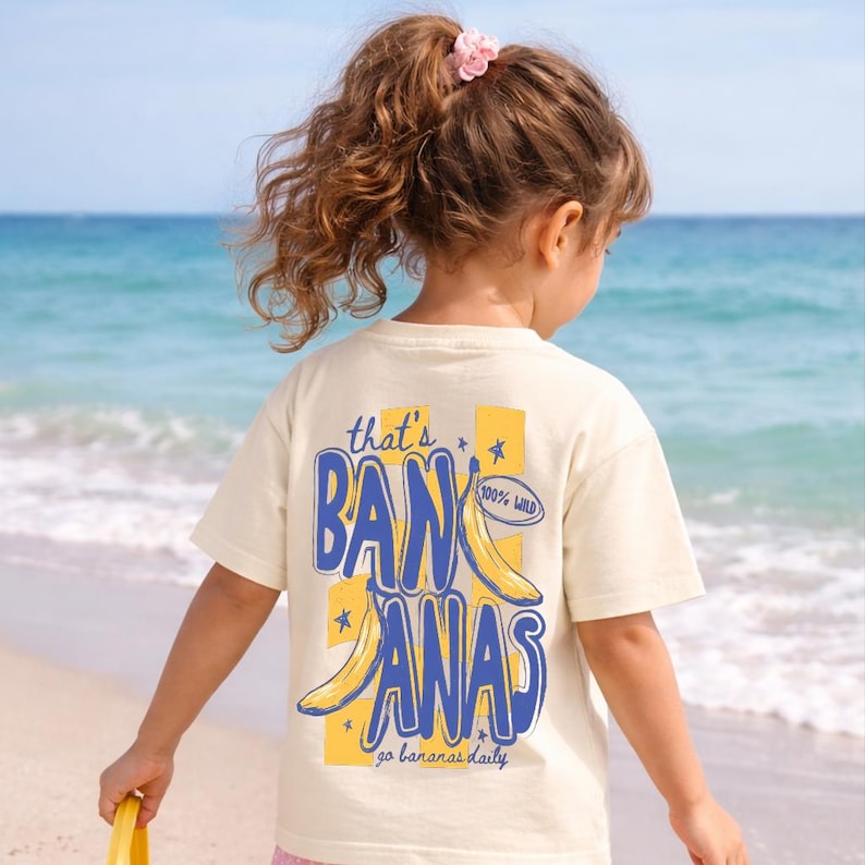 May include: Cream-colored t-shirt with a blue and yellow banana graphic that says "that's BANANAS" and "go bananas daily". The shirt is worn by a child on a beach with the ocean in the background.